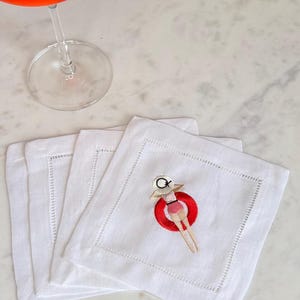 May include: White linen cocktail napkins with a red and white embroidered design of a person floating on a red inner tube. A glass of red cocktail with ice is in the background.