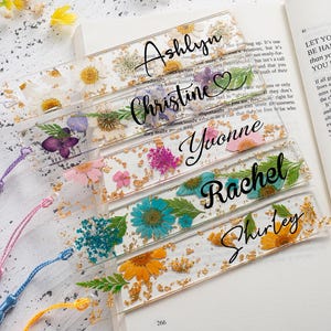 Könnte beinhalten: Transparente Harz-Lesezeichen mit getrockneten Blumen und Goldflocken. Jedes Lesezeichen trägt einen Namen in Schreibschrift: Ashlyn, Christine, Yvonne, Rachel und Shirley. Bunte Quasten in Lila, Rosa, Blau und Gelb sind befestigt.