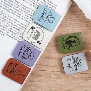 Könnte beinhalten: Fünf verschiedene Leder-Lesezeichen-Designs mit personalisierten Namen und buchbezogenen Illustrationen. Die Lesezeichen sind in verschiedenen Farben, darunter Blau, Grün, Lila, Braun und Grau.