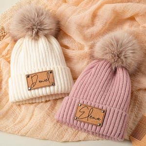 Könnte beinhalten: Zwei gerippte Strickmützen mit Kunstfell-Bommeln. Eine ist elfenbeinfarben mit einem Lederaufnäher mit der Aufschrift "Daniel", die andere ist rosa mit einem Aufnäher mit der Aufschrift "Elena". Eine Holzbürste und ein Kamm sind ebenfalls sichtbar.