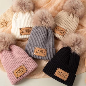 Könnte beinhalten: Eine Auswahl an gerippten Strickmützen in verschiedenen Farben, darunter Weiß, Rosa, Grau, Beige und Schwarz. Jede Mütze hat einen Kunstfell-Bommel und ein rechteckiges Lederetikett mit einem Namen.