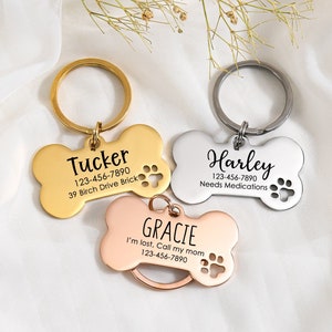May include: Three gold, silver, and rose gold bone-shaped dog tags with engraved text. The tags include the names "Tucker", "Harley", and "Gracie" with phone numbers and additional information.