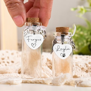 Op de afbeelding: Twee kleine glazen potjes met kurken deksels, elk gevuld met witte vacht en een zilveren hartvormige hanger. De hangers zijn gegraveerd met de namen "Fergus" en "Layla" en hebben een pootprintontwerp.
