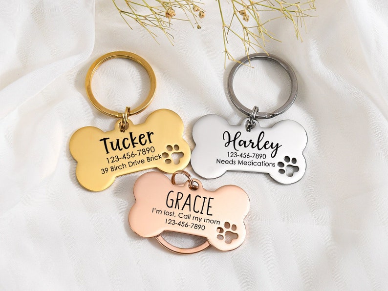 May include: Three gold, silver, and rose gold bone-shaped dog tags with engraved text. The tags include the names "Tucker", "Harley", and "Gracie" with phone numbers and additional information.