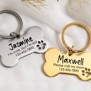 May include: Two bone-shaped dog tags with engraved text and paw prints. The silver tag reads "Jasmine I'm cute, but I'm lost 123-456-7890". The gold tag reads "Maxwell Please call my mom 123-456-7890".