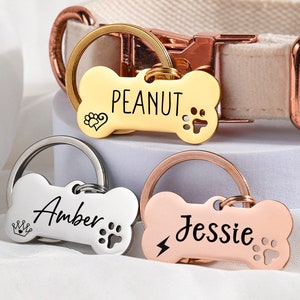 May include: Three dog bone-shaped name tags with different names engraved on them. The tags are attached to key rings. The names on the tags are "Peanut", "Amber", and "Jessie".