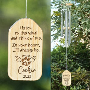 Pode incluir: Carrilhão de vento de madeira com uma mensagem personalizada: "Ouça o vento e pense em mim. No seu coração, eu sempre estarei." O carrilhão tem uma pegada de pata com asas de anjo e o nome "Cookie" e o ano "2023".
