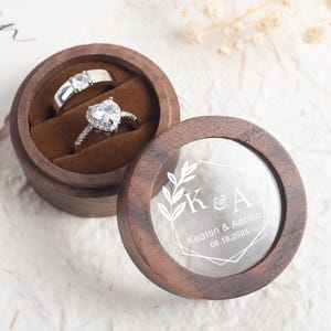 May include: A wooden ring box with a clear lid. The lid has a geometric design with the initials "K & A" and the text "Keaton & Adrian 06.18.2025" engraved on it. The box is open and two rings are inside.