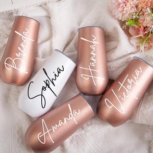 Puede incluir: Cinco vasos personalizados de oro rosa con letras blancas. Los nombres en los vasos son Brenda, Sophia, Hannah, Victoria y Amanda.