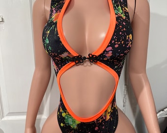 May include: A black bodysuit with a colorful splatter paint design. The bodysuit has a high neckline with a mandarin collar and a cutout at the chest. The neckline is trimmed with orange fabric. The bodysuit has a cutout at the waist and a high leg cut.