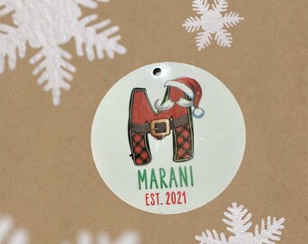 May include: A round Christmas ornament featuring the letter "M" dressed as Santa Claus, with a red and black plaid design. The ornament includes the name "Marani" and the year "EST. 2021" in green text.