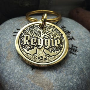 Op de afbeelding: Een goudkleurige, cirkelvormige hondenpenning met de naam "Reggie" in vette letters gegraveerd. De penning heeft een gedetailleerd boomontwerp en een gouden sleutelhanger. De penning ligt op een grijze steen.