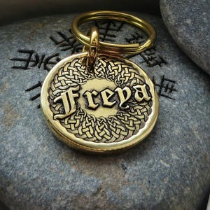 May include: A gold-colored, round pet tag with the name "Freya" in an ornate font, surrounded by a Celtic knot design. The tag is attached to a gold keyring. The background is a gray stone with etched symbols.