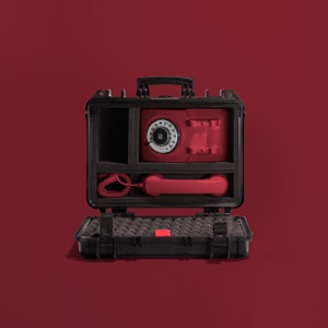 Könnte beinhalten: Ein rotes Wählscheibentelefon und ein kleiner roter Tresor befinden sich in einem offenen, schwarzen Hartschalenkoffer. Der Koffer hat einen Griff und Verschlüsse. Der Hintergrund ist einfarbig rot.