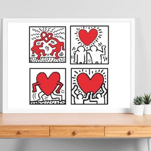 May include: A framed artwork featuring four square panels with Keith Haring-style illustrations. Each panel has a black and white outline with red heart and figure designs. The artwork is displayed on a wooden desk with small potted plants.