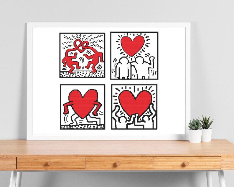 May include: A framed artwork featuring four square panels with Keith Haring-style illustrations. Each panel has a black and white outline with red heart and figure designs. The artwork is displayed on a wooden desk with small potted plants.