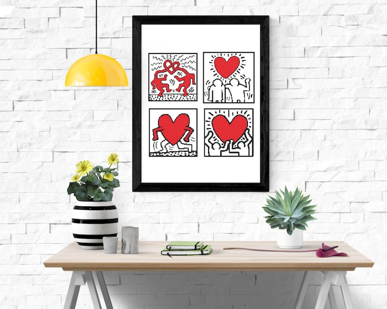 May include: A framed art print featuring four panels with Keith Haring-style illustrations. The artwork includes red figures and hearts on a white background, framed in black. The print is displayed on a light wood table with decorative items.