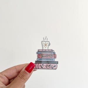 Op de afbeelding: Een sticker van een stapel boeken met een kopje koffie erop. De boeken zijn roze, blauw en bruin met bloemmotieven. Het koffiekopje is wit met een gele zon erin.