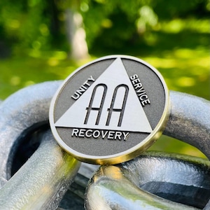 May include: A silver and gold AA medallion with the words "Unity", "Service", and "Recovery" encircling the AA symbol. The medallion is resting on a silver chain, with a blurred green background.