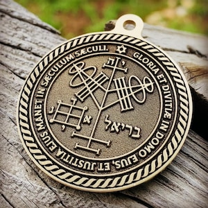Peut inclure: Un pendentif doré avec des symboles et du texte gravés. Le pendentif présente un motif central avec des formes géométriques et du texte hébreu, entouré d'une bordure circulaire avec des inscriptions latines. Une petite boucle est en haut pour suspendre.