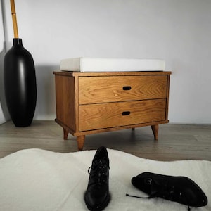 May include: A wooden storage bench with two drawers and a white cushioned top. The bench has a light wood finish and black metal hardware.