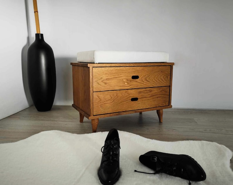 May include: A wooden storage bench with two drawers and a white cushioned top. The bench has a light wood finish and black metal hardware.