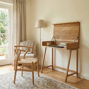 May include: A light oak writing desk with a hinged top, a keyboard, and a mug. A wooden chair with a woven seat and a floor lamp are nearby. The room has a neutral colour palette with a rug.