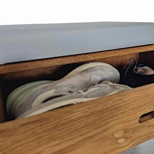 May include: A wooden shoe storage bench with a light gray cushioned top and a drawer that is open, revealing a pair of white sneakers and a pair of black shoes.