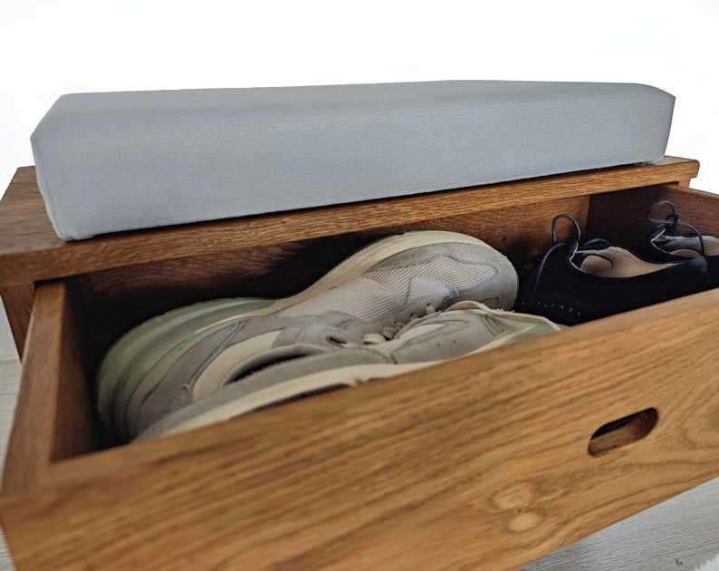 May include: A wooden shoe storage bench with a light gray cushioned top and a drawer that is open, revealing a pair of white sneakers and a pair of black shoes.