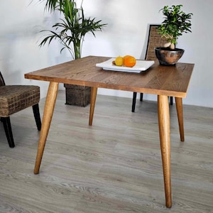 Pode incluir: Mesa de jantar quadrada de madeira com acabamento natural e pernas anguladas. Um prato retangular branco com um limão e uma laranja está sobre a mesa. Uma pequena árvore bonsai em um vaso escuro também está sobre a mesa.