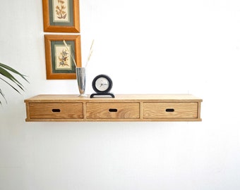 Könnte beinhalten: Ein schwebendes Regal aus hellem Holz mit drei Schubladen. Das Regal ist an einer weißen Wand montiert und hat eine Uhr und eine Vase mit getrockneten Blumen darauf.