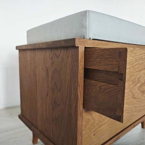 May include: A wooden storage bench with a light gray cushioned top and a drawer that is partially open. The bench has a dark wood stain and is made of solid wood.