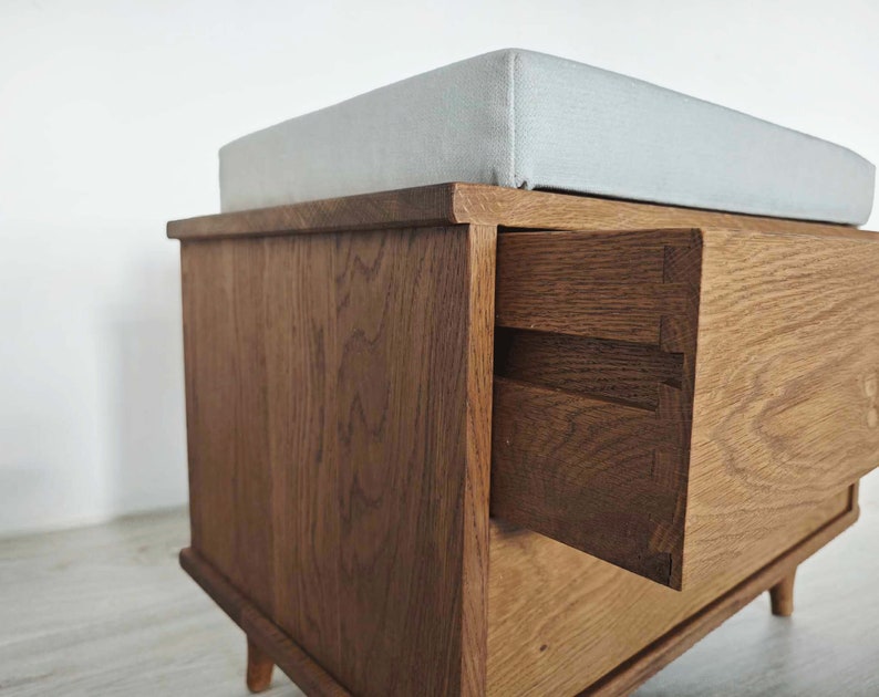 May include: A wooden storage bench with a light gray cushioned top and a drawer that is partially open. The bench has a dark wood stain and is made of solid wood.