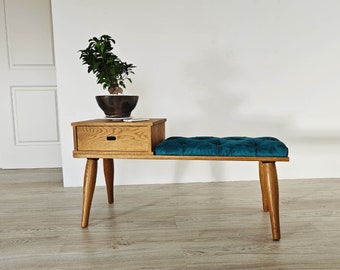 Op de afbeelding: Een houten bank met een turquoise fluwelen kussen en een lade eronder. De bank heeft vier poten en is gemaakt van licht hout. Een kleine plant in een pot staat op de lade.