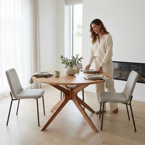 Op de afbeelding: Een ronde eettafel van licht hout, gedekt met borden, kopjes en een vaas met groen. Twee grijs gestoffeerde stoelen met zwarte metalen poten staan rond de tafel. Een persoon dekt de tafel in een lichte, moderne eetkamer.