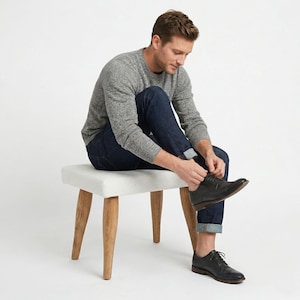 Könnte beinhalten: Ein grauer Pullover und dunkelblaue Jeans. Die Person sitzt auf einem kleinen, weiß gepolsterten Hocker mit Holzbeinen und bindet die Schnürsenkel eines schwarzen Lederschuhs. Der Hocker ist etwa 46 cm breit.