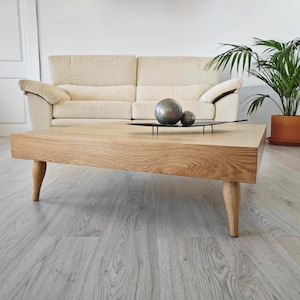 Peut inclure: Une table basse en bois clair avec un plateau carré et quatre pieds. La table est dans un salon avec un canapé blanc et une plante en pot en arrière-plan.