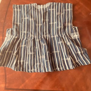May include: A sleeveless top featuring a vertical striped pattern in shades of dark blue, light blue, and white. The garment has a gathered, flared hemline and white embroidered detailing around the neckline and the side arm openings.
