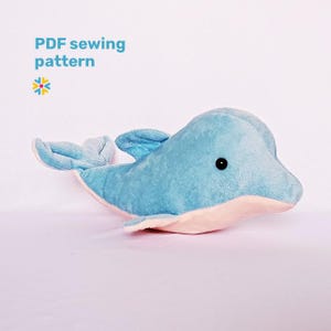 Op de afbeelding: Een zacht, blauw dolfijnknuffel met een lichtroze buik en zwarte ogen. De tekst "PDF sewing pattern" staat in de linkerbovenhoek. De dolfijn heeft een getextureerd uiterlijk, wat suggereert dat hij van stof is gemaakt.