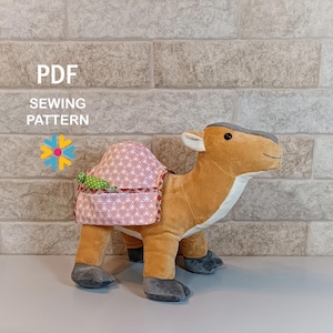 May include: A plush camel toy with a pink and white geometric patterned saddle. The camel is tan with gray accents on its feet and head. The image includes the text "PDF SEWING PATTERN".