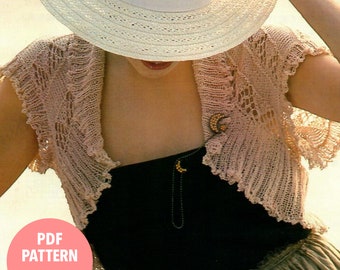 May include: A pale pink, open-front knitted shrug with a delicate, lacy design. The shrug features short sleeves and a scalloped edge. A gold-toned crescent moon brooch is pinned on the front. The image also includes a white sun hat and the text "PDF PATTERN".