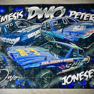 May include: A jigsaw puzzle depicting three blue racing cars, each with sponsor logos and names like "NEMESIS", "DWO", and "PETERS" in white. The background is dark blue and black with abstract patterns.