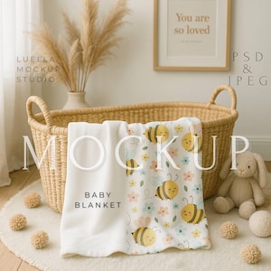 May include: A white baby blanket with a bee and flower pattern, folded in a wicker basket. The blanket has the words "Baby Blanket" printed on it. A stuffed bunny and decorative balls are nearby.