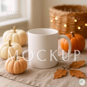 Könnte beinhalten: Eine weiße Keramiktasse steht im Mittelpunkt dieses herbstlichen Stilllebens. Miniaturkürbisse in Orange- und Cremetönen umgeben die Tasse, mit Herbstblättern und einem Weidenkorb im Hintergrund. Das Wort "MOCKUP" ist auf die Tasse gelegt.