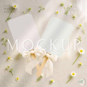 May include: Two white rectangular mockups with wooden handles and cream-colored bows. The word "MOCKUP" is overlaid on the image. Daisies and small white flowers are scattered around the mockups, creating a soft, natural aesthetic.