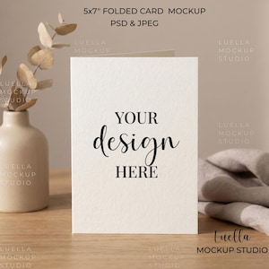May include: A 5x7 inch folded card mockup with the text "YOUR design HERE" in black script. The card is on a wooden surface, with a vase of dried flowers and a folded cloth in the background. The mockup is labeled "PSD & JPEG".