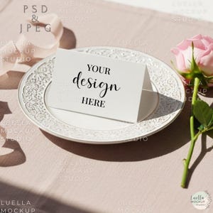 May include: A white card with the words "YOUR design HERE" sits on a white, ornate plate. A pink rose and greenery are to the right. The background is a light pink fabric.