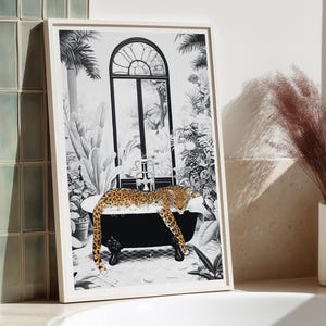 May include: A framed black and white art print featuring a leopard resting in a clawfoot bathtub. The scene includes a large arched window, lush tropical plants, and a tiled floor. The leopard has a spotted coat and is positioned inside the tub.