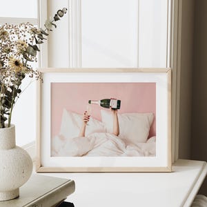 May include: A framed print shows a person pouring champagne into a glass while in bed. The champagne bottle is green with a white label. The background is a light pink wall and white bedding.