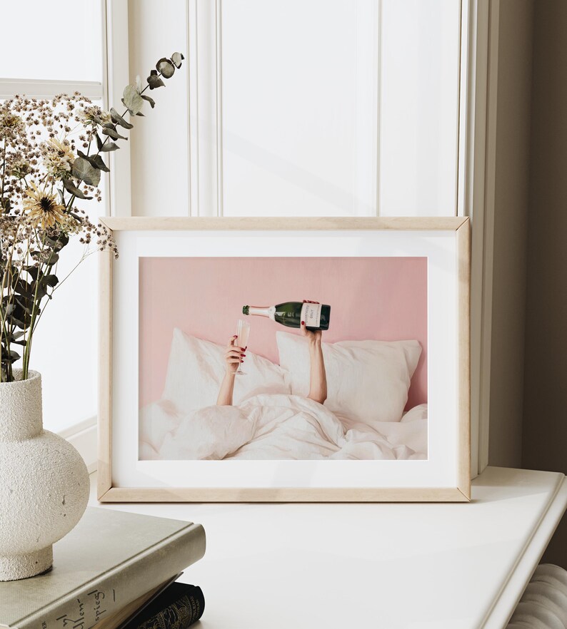 May include: A framed print shows a person pouring champagne into a glass while in bed. The champagne bottle is green with a white label. The background is a light pink wall and white bedding.